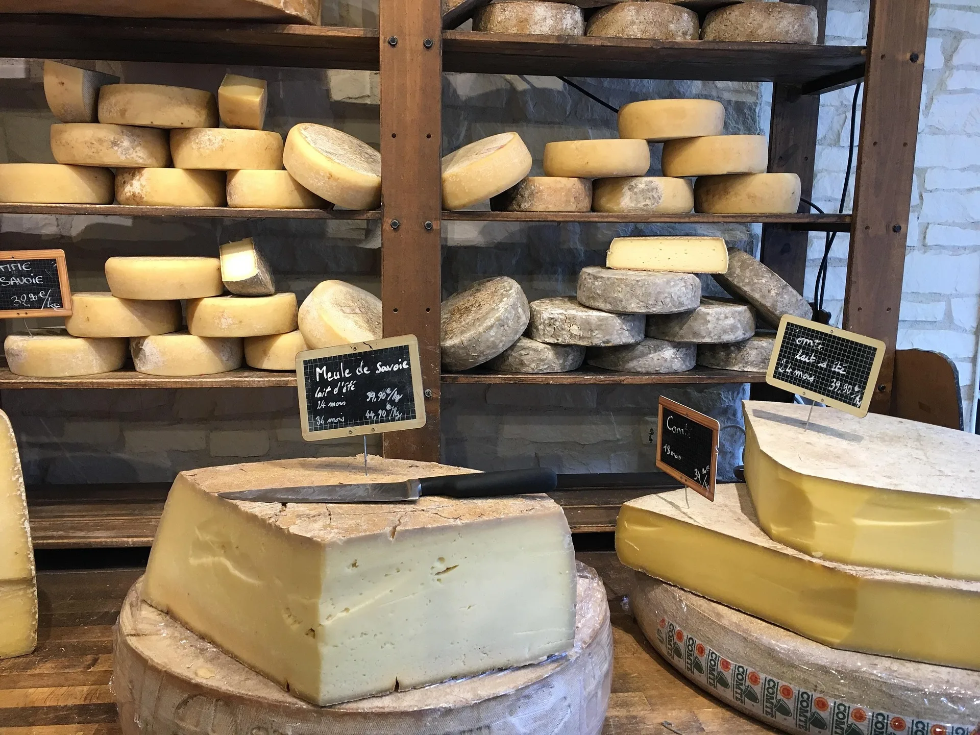 Fromages artisanaux sur étagères en bois dans une fromagerie, avec meules affinées, morceaux découpés et étiquettes de prix visibles.