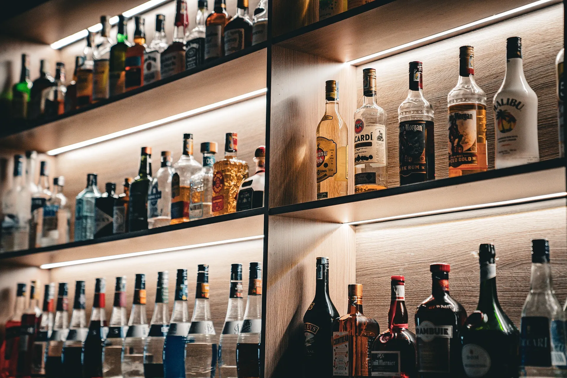 Étagères rétroéclairées avec bouteilles d’alcool variées (rhum, vodka, liqueurs) dans un bar moderne.